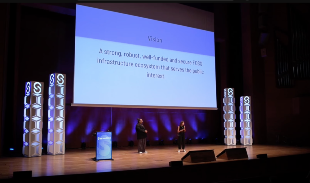 Tara Tarakiyee und Fiona Krakenbürger präsentieren auf einer großen Bühne auf der Open Source Summit Europe in Bilbao. Auf der Leinwand steht "Vision: A strong, robust, well-funded and secure FOSS infrastructure ecosystem that serves the public interest."